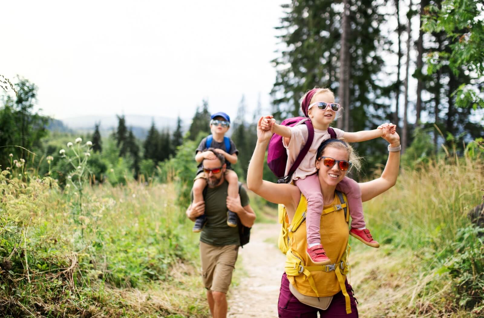 A family hiking in a forest, with two adults carrying children on their shoulders, all wearing sunglasses and backpacks.