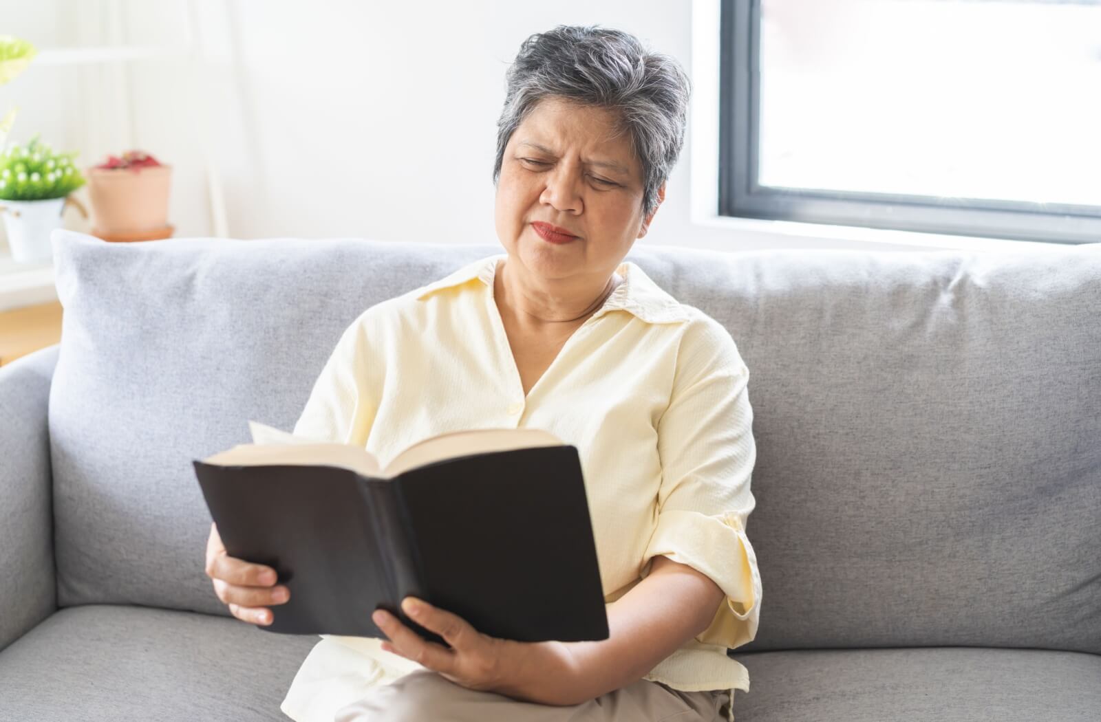 A woman sitting on a couch and straining to read a book held at arm's length.